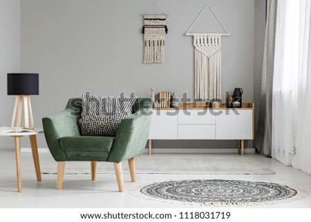 Real photo of a green armchair standing in a bright, natural living room interior with macrame hanging on gray wall above white cupboard