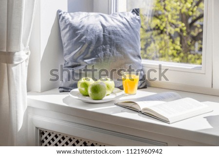 Grey pillow, book, apples and orange juice on window sill in bright interior. Real photo