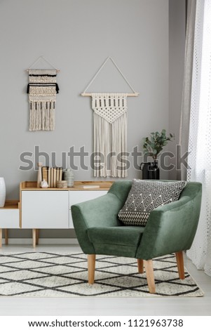 Real photo of a boho living room interior with macrame hanging on gray wall behind a comfy, green armchair with a cushion