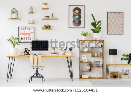 Real photo of a desk with a computer screen, ornaments and a plant standing next to shelves with plants in workplace with posters and small shelves on white wall