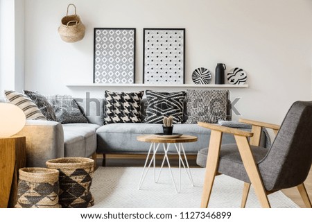 Real photo of a small table standing between a sofa with pillows and an armchair on a white rug in boho living room interior with posters on a shelf