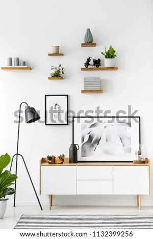 Black lamp next to cupboard against white wall with posters in simple flat interior. Real photo