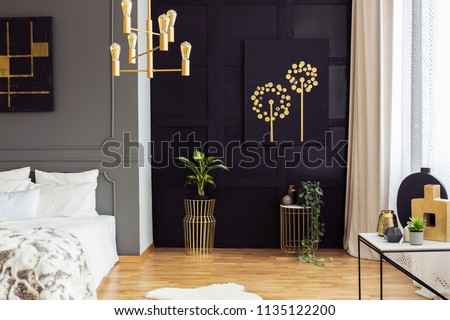 Black and gold poster above plants in bedroom interior with white pillows on bed. Real photo