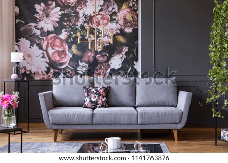 Grey lounge with patterned cushion in real photo of dark living room interior with floral wallpaper, molding on the wall and gold lamp