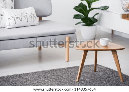 Cup on a wooden coffee table and blurry background with graphic pillows on a gray sofa in a white living room interior