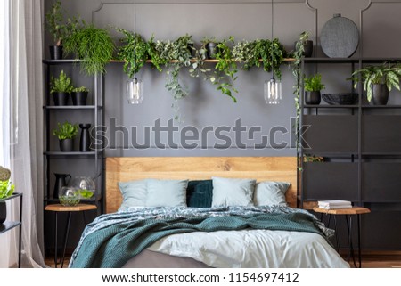 Green blanket and grey pillows on wooden bed in floral bedroom interior with plants. Real photo