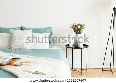 Close-up of a bed with pale sage green and white linen, pillows and a blanket in a sunny bedroom interior. A round black metal side table with vases and flowers beside the bed. Real photo