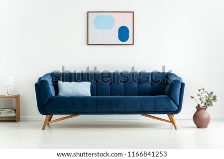 Plant next to blue settee with cushion in white minimal living room interior with poster. Real photo