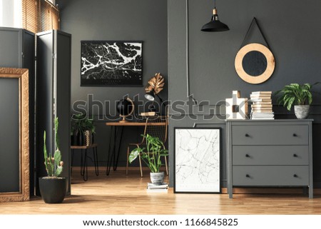 Black map on grey wall in dark living room interior with plants and poster. Real photo