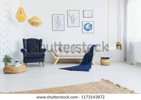Real photo of a bright living room interior with a comfy armchair, sofa, wicker lamps and botanical graphics on the wall