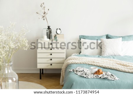 A bright bedroom interior with sage green and white bedding, pillows on bed and a drawer nightstand. Real photo.