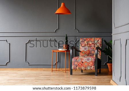 Bright orange ceiling light above a boho style armchair in an elegant living room interior with molding on gray walls and copy space place for a chair. Real photo.