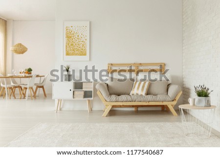 Gold poster above white cabinet next to beige wooden settee in loft interior with table. Real photo