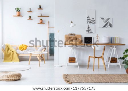 Chair at wooden desk and pouf in white open space interior with posters and table near sofa. Real photo