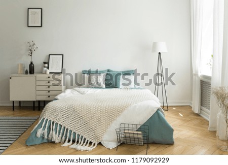 A big comfortable bed with pale sage green and white linen, pillows and blanket in a woman\'s bright bedroom interior with windows. Real photo.