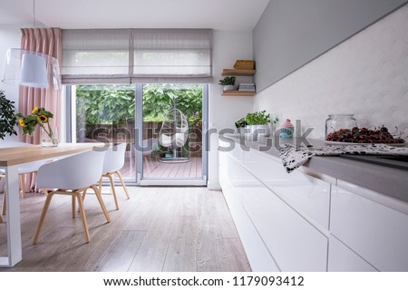 Hanging chair on a wooden patio of a modern house with white kitchen interior. Real photo of cherries on gray counter and sunflowers on wooden dining table