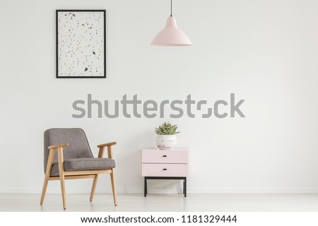Poster above grey wooden armchair next to cabinet with plant in white flat interior with lamp. Real photo