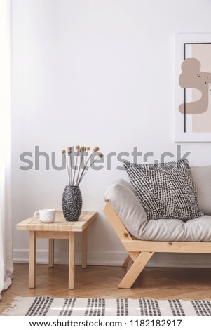 Flowers on wooden table next to beige couch with cushion in flat interior with poster. Real photo