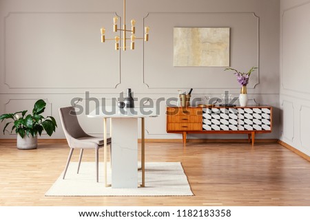 Chair at table under gold lamp in living room interior with poster above cupboard with plant. Real photo