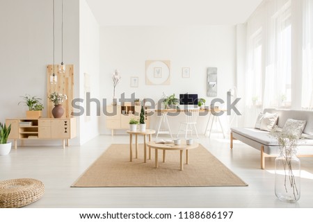 Wooden tables on carpet next to grey sofa in white apartment interior with plants and pouf. Real photo