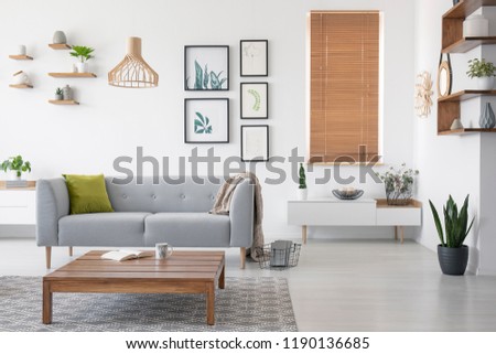 Wooden table in front of grey settee in natural living room interior with blinds and gallery. Real photo