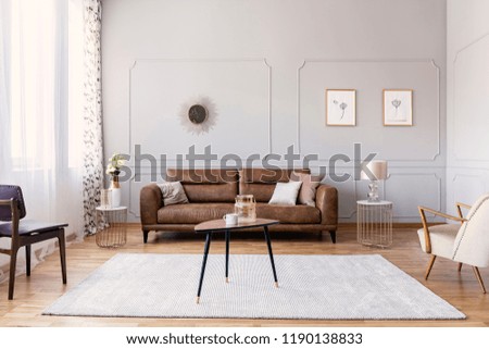 Coffee table with vase and mug in the middle of elegant living room interior with comfortable leather sofa, stylish purple chair and armchair
