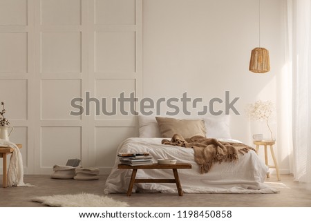 Natural blanket on white bed in simple bedroom interior with fur next to wooden stool. Real photo