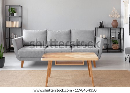 Wooden table on carpet in front of grey sofa in minimal living room interior with plant. Real photo