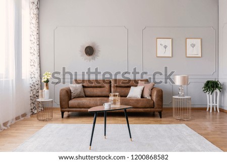 Wooden table on carpet in front of leather sofa in grey living room interior with posters. Real photo