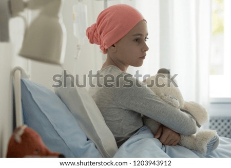 Sick child with cancer sitting in hospital bed holding teddy bear