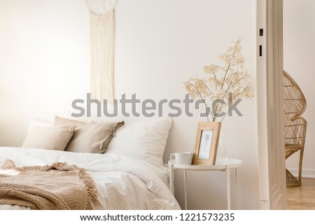 Bedside table with mug and framed picture next to comfortable king size bed with linen bedding and beige blanket, real photo