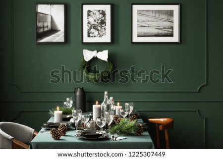 Christmas decor in stylish dining room interior with communal table with plates, wine glasses and candles, real photo with christmas wreath and posters in black frames on empty green wall