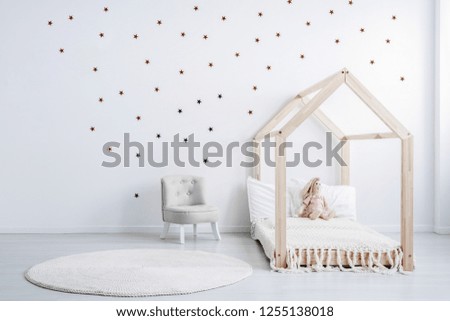 Pastel grey armchair next to wooden house shape bed with toy and blanket, copy space and golden stars on empty white wall, round white carpet on floor