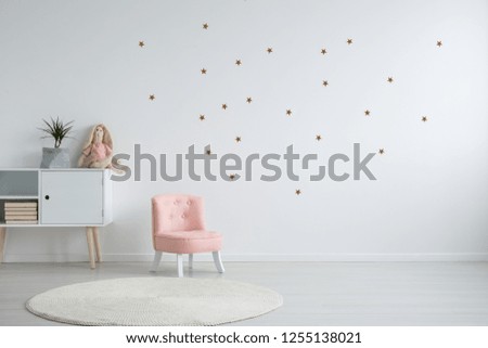 Pastel pink armchair next to wooden cabinet with books, toy and green plant in grey material pot, copy space and golder stars stickers on empty white wall, round carpet on the floor