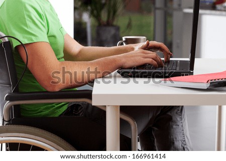 Young man on wheelchair writing on laptop and drinking coffee