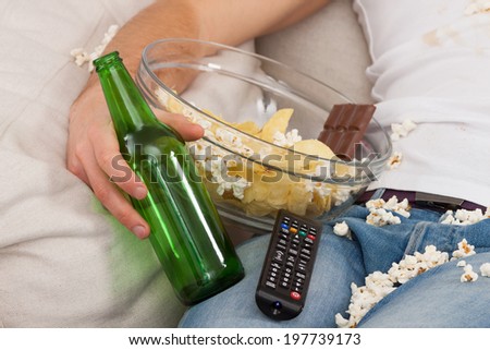 A closeup of a man with a bowl of junk food, beer and a remote control