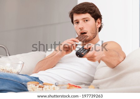 A man eating a chocolate bar holding a remote control on a couch