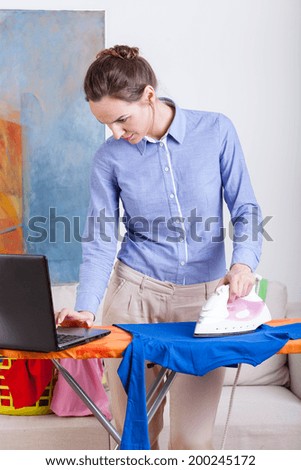 Beautiful young woman ironing clothes and checking email on her laptop