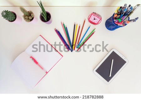 Top view of open exercise book, pencil crayons and tablet on white desk