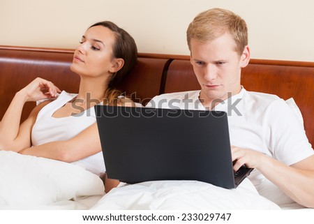 A man surfing on his laptop and his bored woman in bed