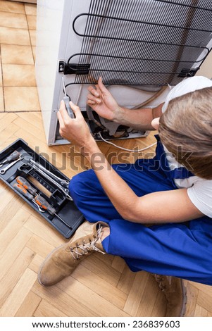 A repairman sitting on the floor fixing a fridge with special tools