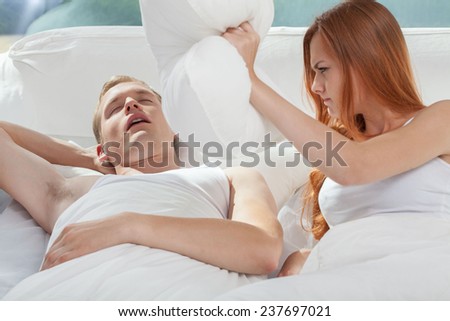 Young snoring man in bed and his angry woman holding the pillow