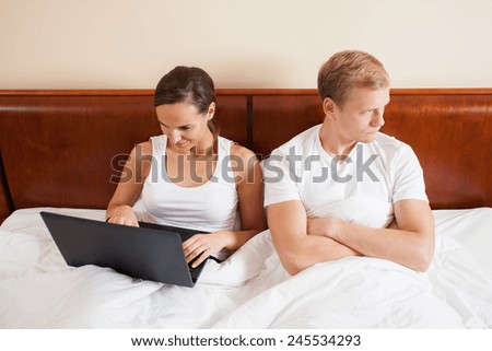 A woman surfing on her laptop and her bored husband in bed