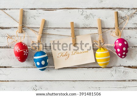 Decorative easter eggs hanging on thin rope, card with wishes in portuguese, light planks in the background/Have a peaceful easter!/Happy Easter
