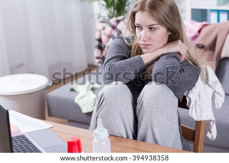 Sad and tired woman with PPD working beside table, looking on laptop, sitting in messy room