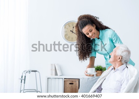 Pretty young caregiver serving afternoon cup of tea to older happy man