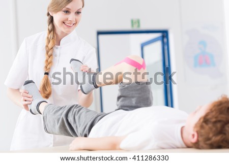 Young pretty smiling physiotherapist exercising with little boy after knee injury