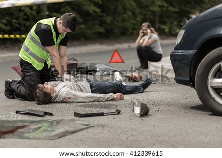 Paramedic resuscitating on street car accident victim, in the background woman talking on phone.