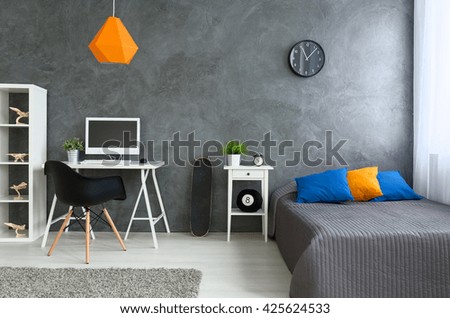 Bedroom with grey walls and grey bed with  colorful pillows and orange lamp. By the wall skate board and desk with computer