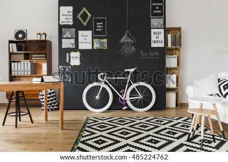 Modern designed room with a black wall with posters on, with wooden table, racks and coffee table, mosaic pattern carpet and white fasionable bicycle